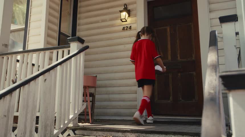 A young girl in a soccer uniform excitedly runs up her front porch steps and slides a note under the door