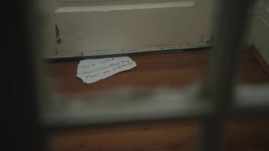 A handwritten invitation to a neighborhood block party is slid under a wooden door onto a hardwood floor