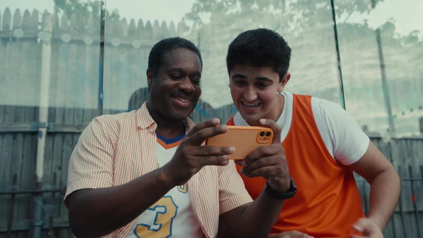 Two diverse friends in basketball jerseys celebrate excitedly after watching a video on a smartphone, giving each other a high-five and a hug