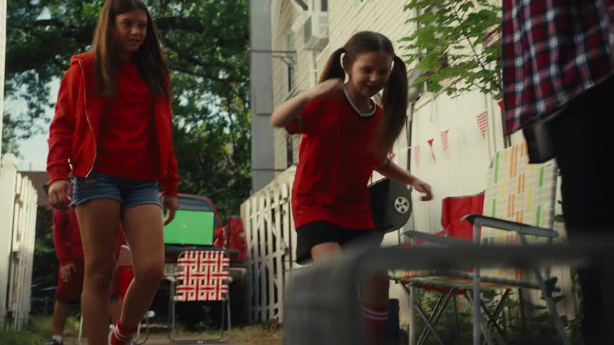 A group of friends and family wearing red celebrate joyfully in slow motion after a young girl scores a goal during a backyard soccer game