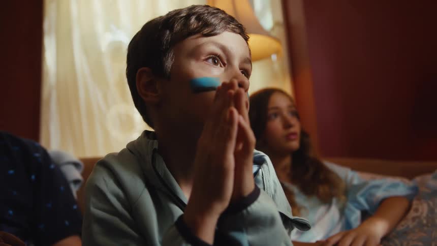 A nervous young boy with blue face paint watches a game with his family and then explodes with joy, kicking and cheering in celebration