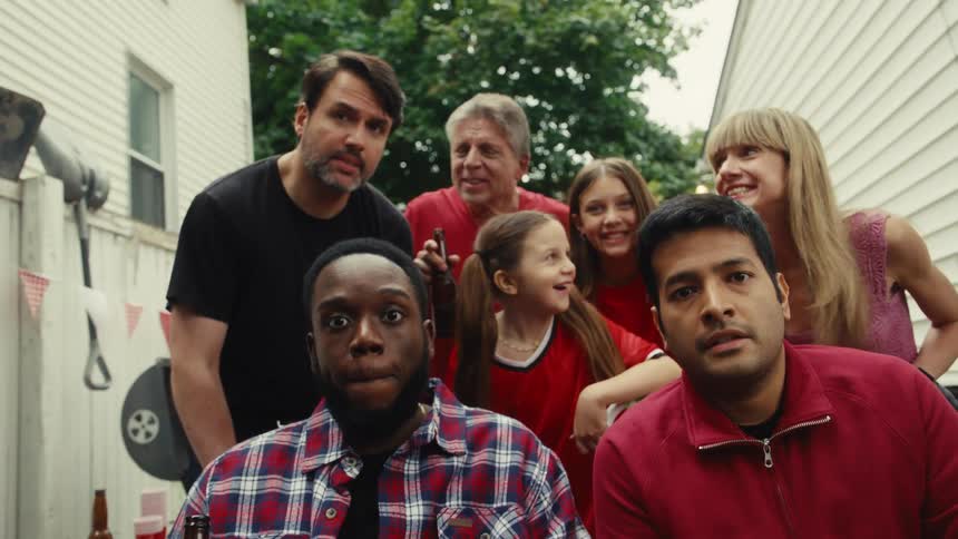 A diverse group of friends and family watch an intense game together in a backyard, erupting in celebration when their team scores