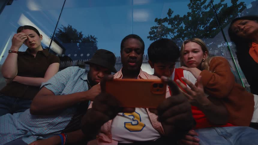 A diverse group of friends huddle around a smartphone at a bus stop, their suspense turning to disappointment as they watch their team lose the game