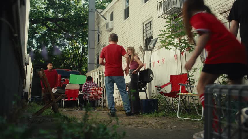 A family and friends enjoy a backyard tailgate party while a young girl in a soccer uniform practices with a ball in slow motion