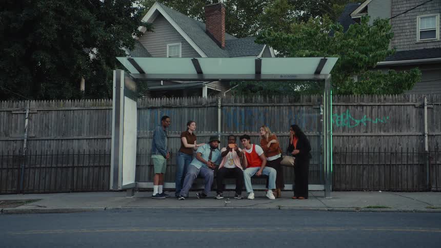 A diverse group of young people waits at a bus stop in a residential neighborhood as traffic passes in the foreground