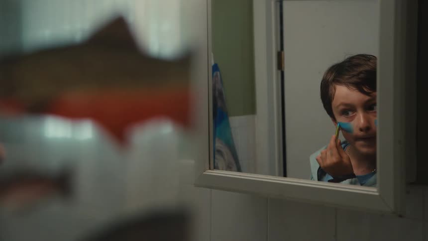 A young boy's reflection in a bathroom mirror shows him seriously applying colorful face paint with a brush