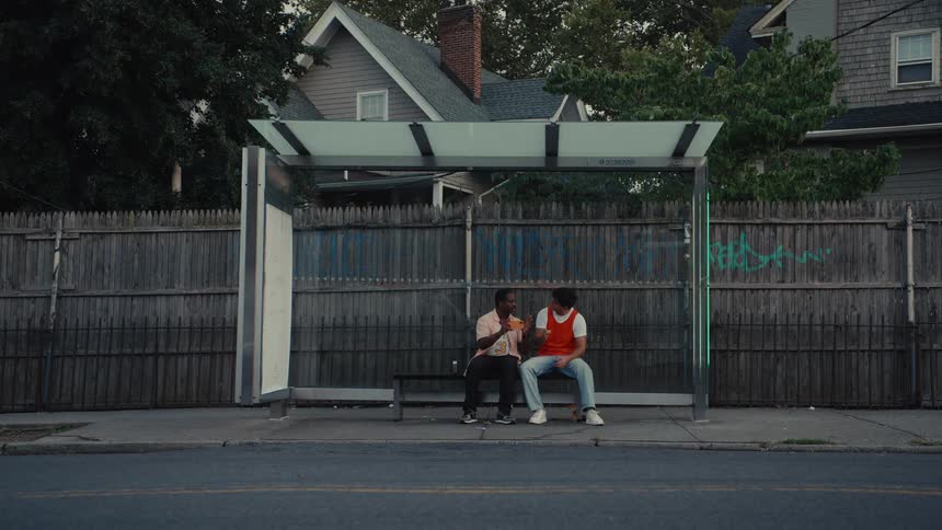 A wide, static shot captures two young friends talking at a bus stop while traffic streams past on a suburban street