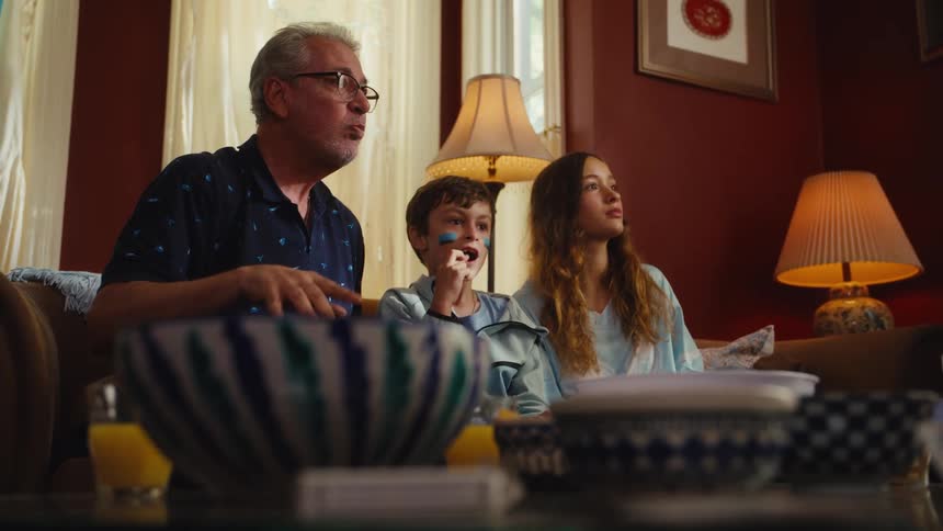 In slow motion, a grandfather and his two grandchildren eat snacks and get excited while watching a sports match at home