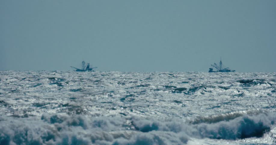 Two fishing boats are silhouetted on the horizon of a vast, turbulent ocean with large waves in the foreground