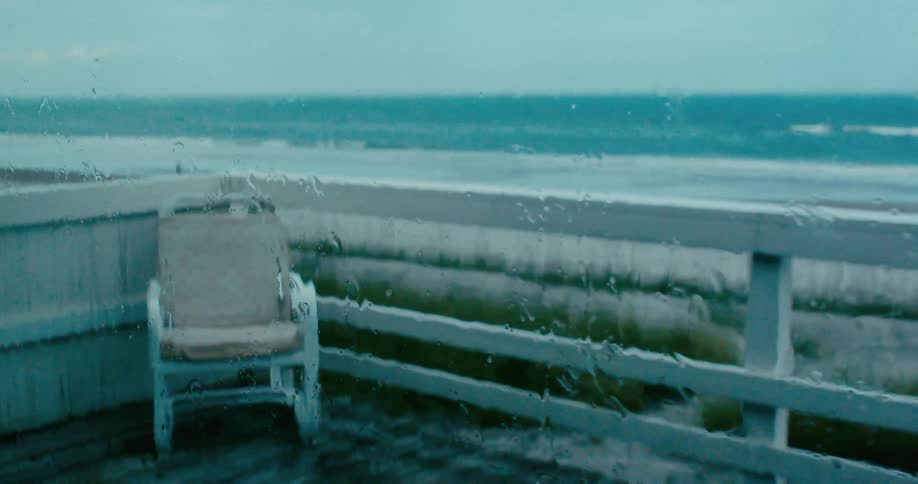 Rain streams down a windowpane overlooking an empty deck chair and the ocean during a storm