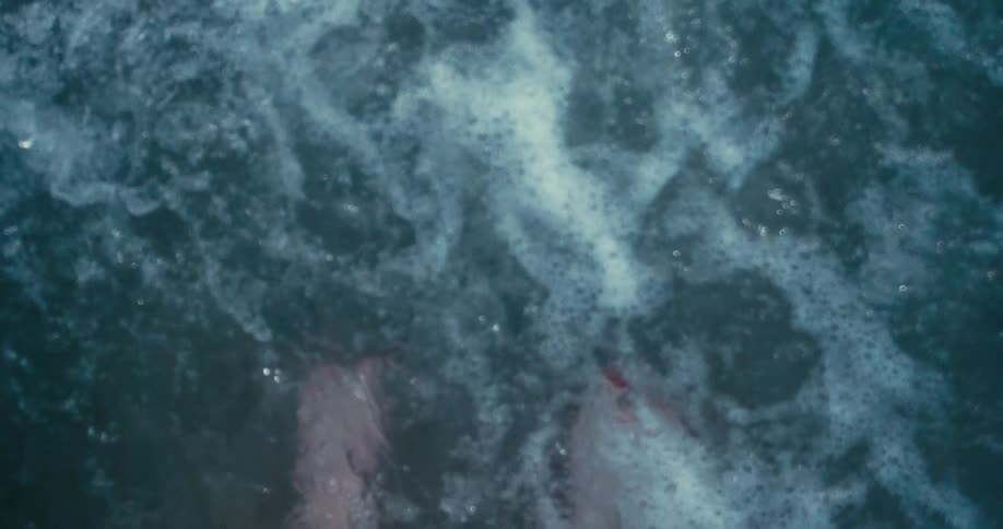 A personal perspective of a person's feet with red painted toenails being washed over by foamy ocean waves in super slow motion