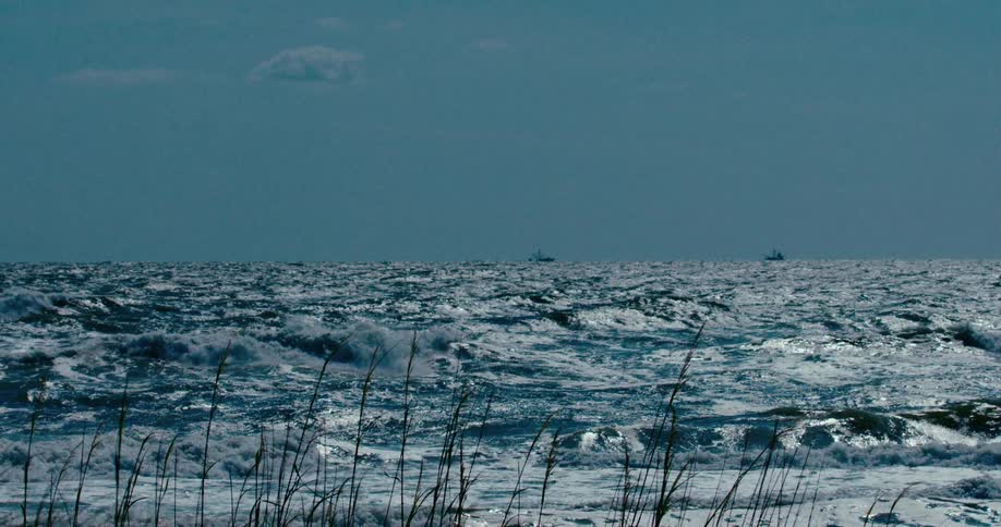 A dramatic slow-motion shot captures the churning, whitecapped waves of a stormy sea with two ships visible on the distant horizon