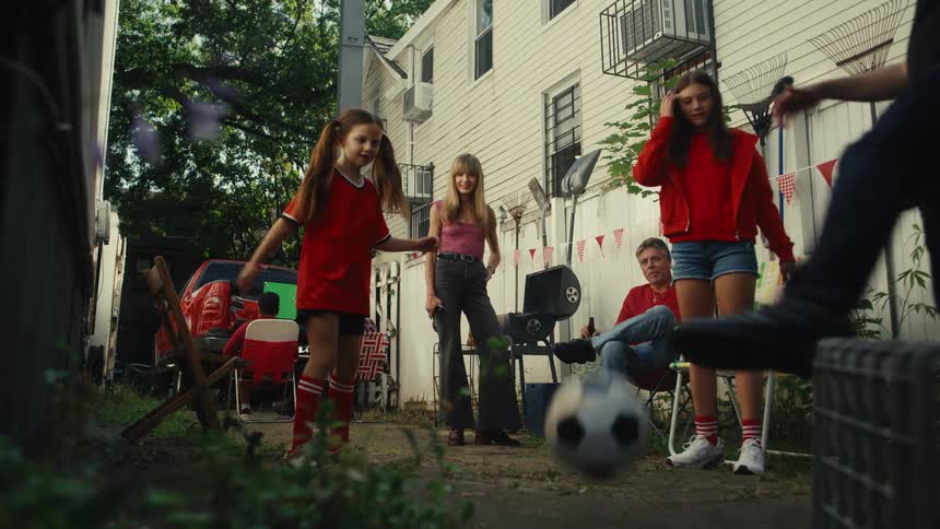 A family and their friends enjoy a backyard gathering, playing soccer together in slow motion between their houses