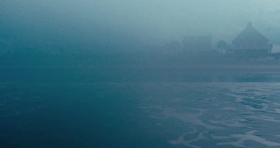 Calm ocean water in the foreground with an eerie, fog-covered coastal town in the background
