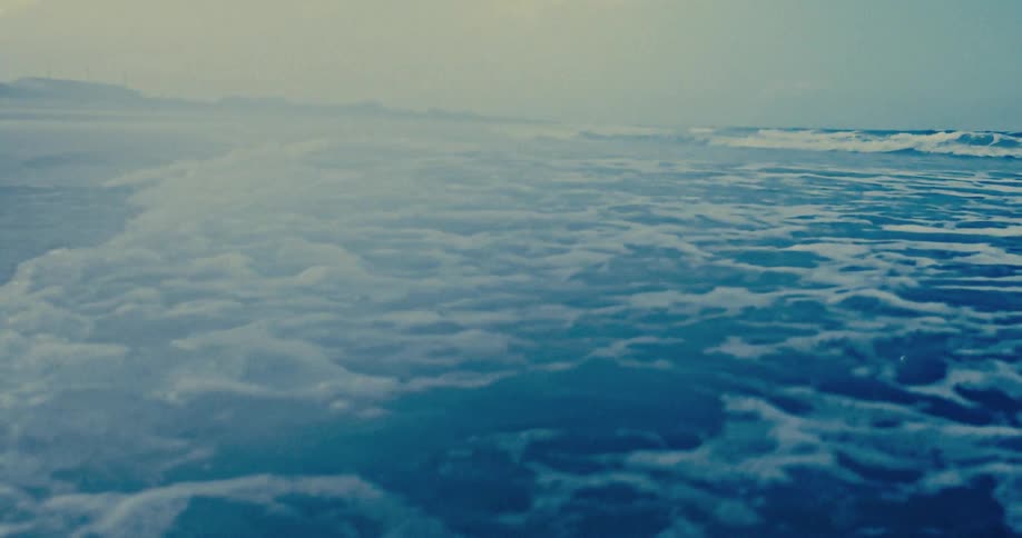 Slow-motion shot of a gentle ocean wave with white foam washing over the blue sea toward a misty shoreline