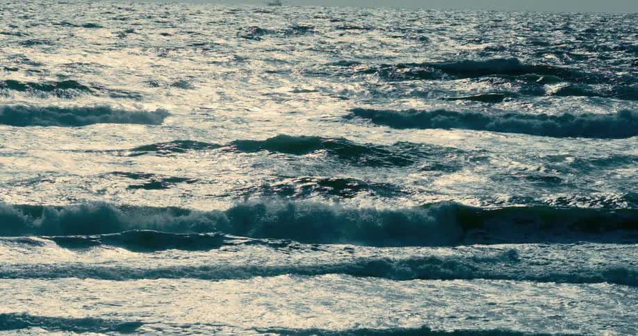 Sunlight glints off the surface of choppy ocean waves moving in slow motion with a ship on the horizon