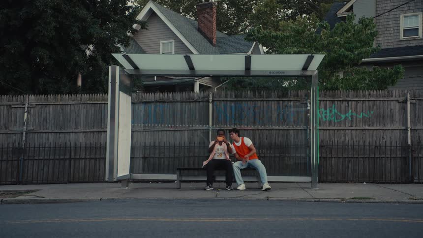 Two young men sit together at a neighborhood bus stop while one eats a sandwich and traffic drives by