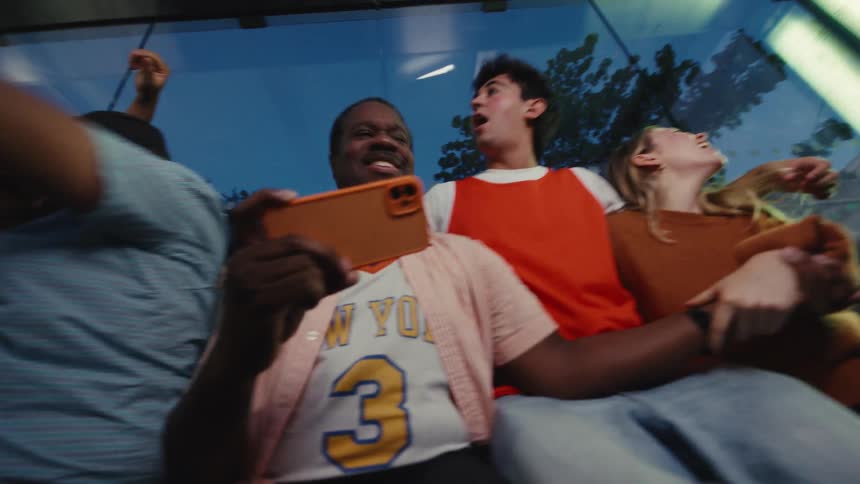 A diverse group of sports fans huddled together at a bus stop at night explode with joy as they watch their team win on a smartphone in slow motion