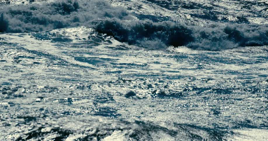 A powerful, hypnotic slow-motion shot of sunlight glinting off the dark, churning surface of ocean waves