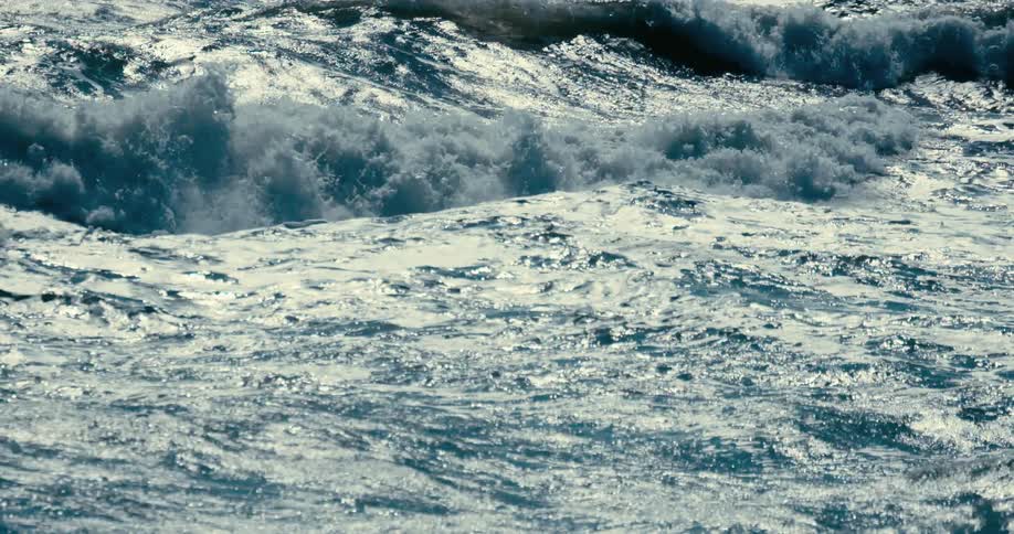 Sunlight reflects off the surface of turbulent, foamy ocean waves in slow motion