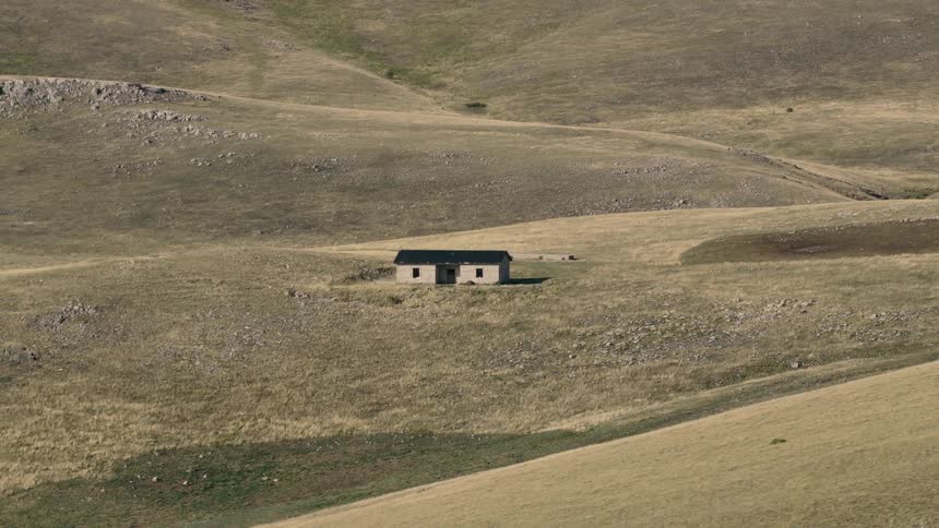 A slow zoom out from a single, isolated stone house situated in the middle of a vast and desolate hilly grassland