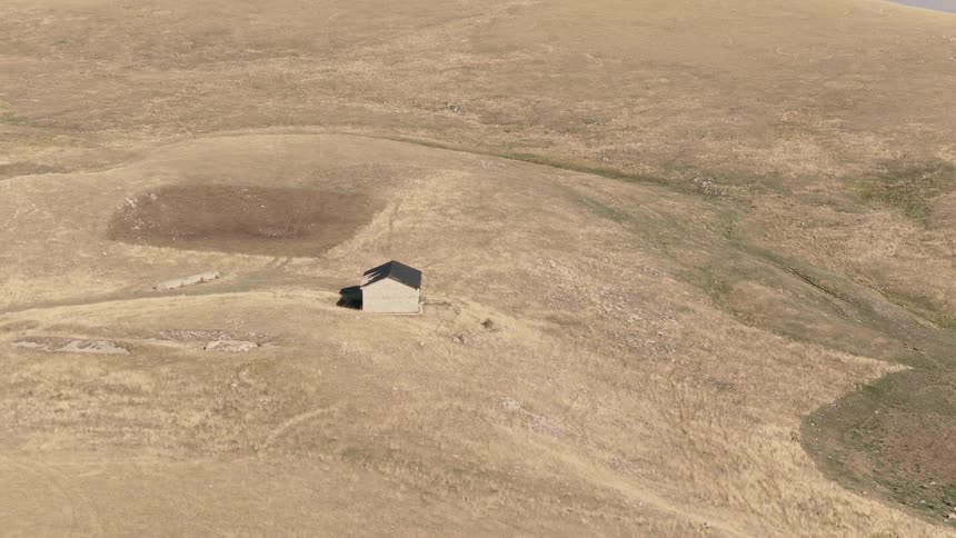 Slow aerial pullback reveals an isolated stone cabin nestled in the vast, arid, golden hills of a remote mountain range