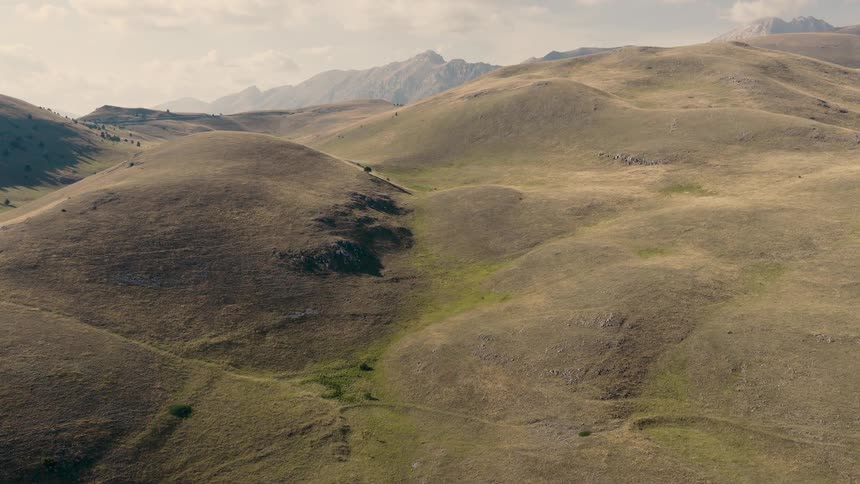 An expansive aerial shot slowly moves over undulating grassy hills with a distant mountain range on the horizon