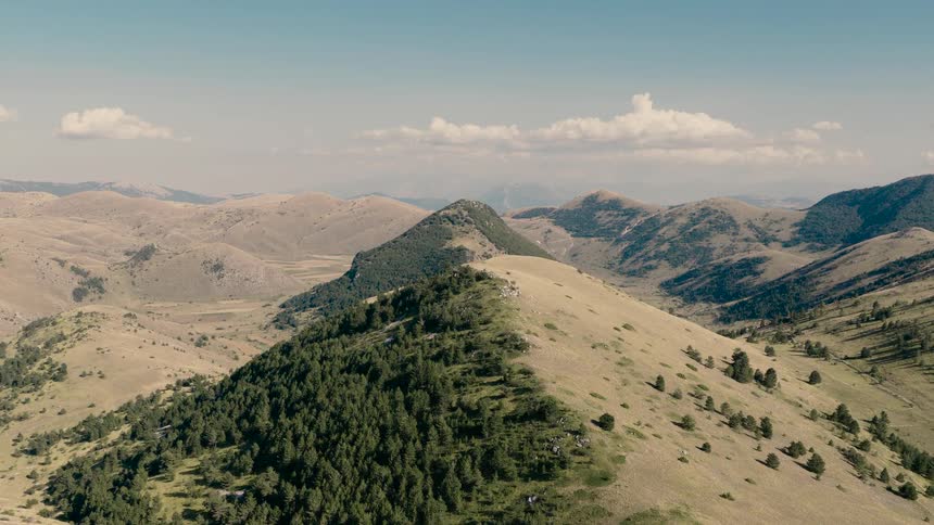 Epic aerial shot slowly reveals a vast, rolling mountain range with a prominent forested ridge under a beautiful blue sky