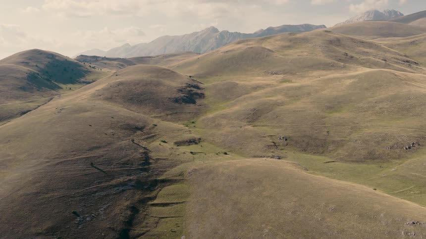 A slow aerial flight over vast, rolling hills of green and brown grass with a rugged mountain range in the distance