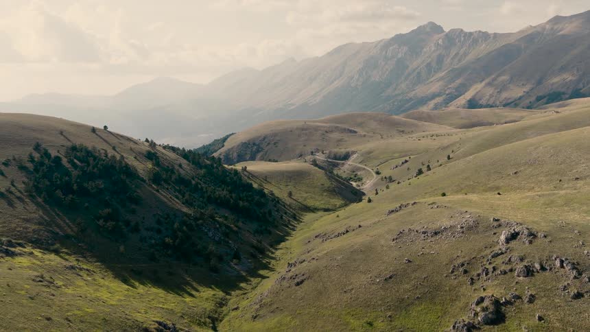 A sweeping aerial view reveals a vast, sunlit valley of rolling green hills leading towards a majestic mountain range in the distance