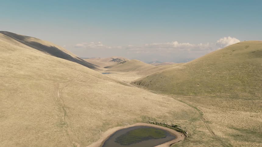 Aerial drone footage reveals a secluded pond in a vast, arid valley where a herd of sheep gathers to drink