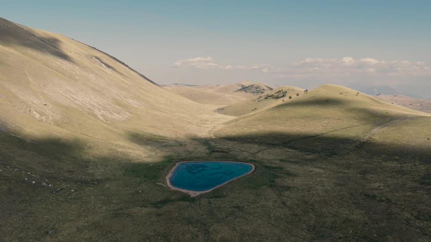 An aerial journey over a vast, sun-drenched mountain valley reveals a hidden, pristine blue lake
