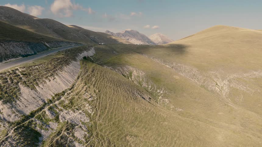 A stunning aerial view follows vehicles on a scenic mountain pass winding through vast, rolling green hills