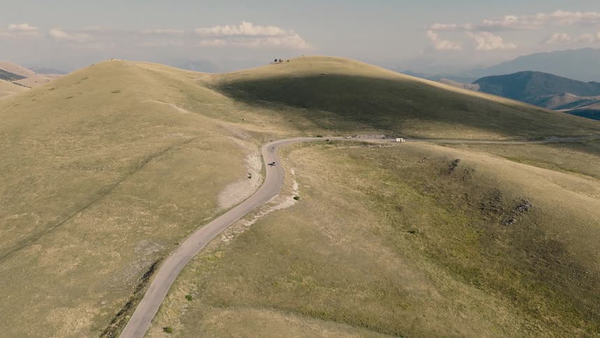 Aerial view of cars and a cyclist traveling along a winding road through a vast, sunlit mountain landscape