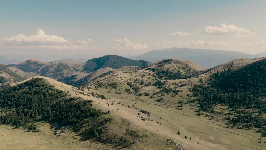 A slow aerial dolly shot reveals the vast expanse of rolling green hills and mountains under a partly cloudy sky