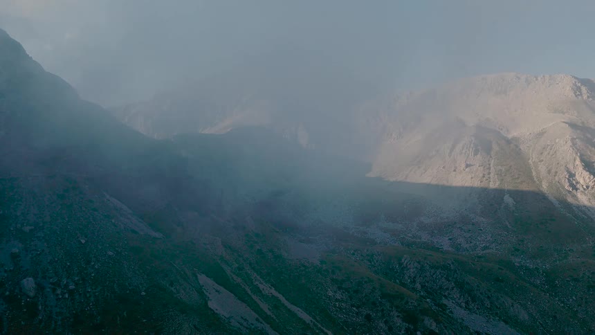 An aerial drone shot flies through thick morning fog, slowly revealing the majestic and rocky mountain range of Gran Sasso in Italy
