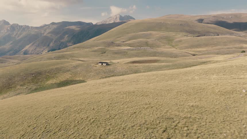 An aerial view slowly advances over vast, rolling green hills towards a solitary, remote house nestled in the mountains