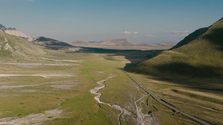 Expansive aerial shot flying through a stunning mountain valley, revealing a dry riverbed and a road below on a sunny day