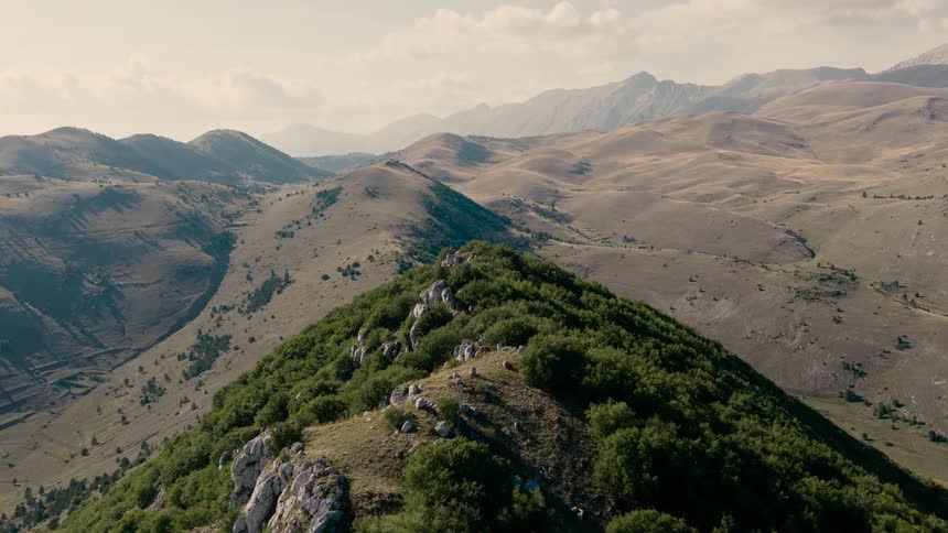 A sweeping aerial view follows a lush green mountain ridgeline, with a vast, sun-drenched valley and distant peaks stretching into the hazy horizon