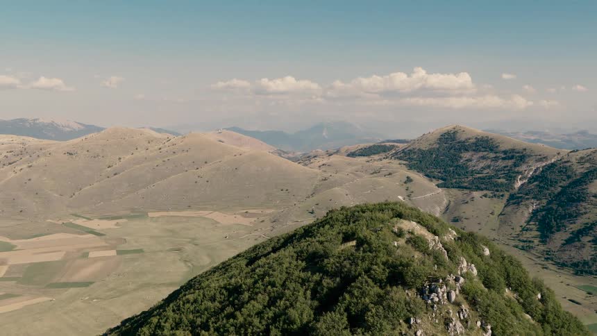 An aerial shot slowly ascends over a tree-covered mountain peak, revealing a vast and scenic landscape of rolling hills and valleys under a blue sky