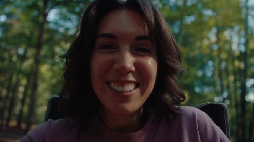 A joyful woman sits in a camping chair and smiles at the camera, relaxing at her campsite in the sunny forest