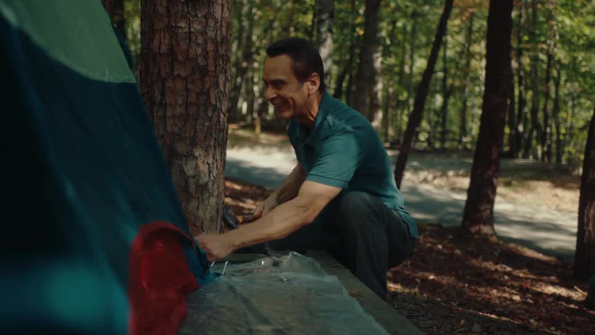 A happy man hammers tent stakes into the ground while setting up camp in a beautiful, sunny forest