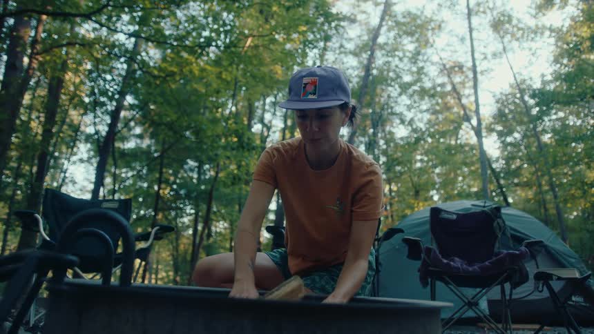 A woman carefully arranges firewood in a fire pit at her campsite in the forest