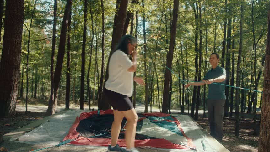 A couple works together to set up their tent at a sunny campsite in the forest
