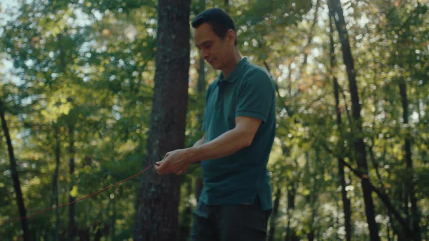 A man carefully ties a knot in a red rope while enjoying a sunny day in the forest