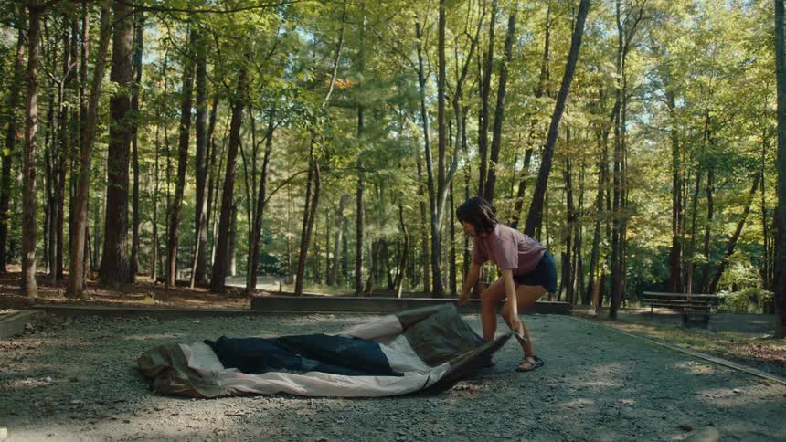 A woman unfolds a tent in slow motion at a beautiful, sunny campsite surrounded by trees