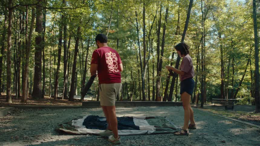A couple works together to set up their tent at a sunny campsite in the forest
