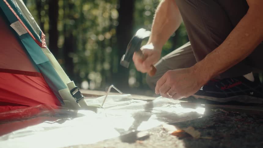 A man hammering a tent stake into the ground while setting up camp in a sunny forest