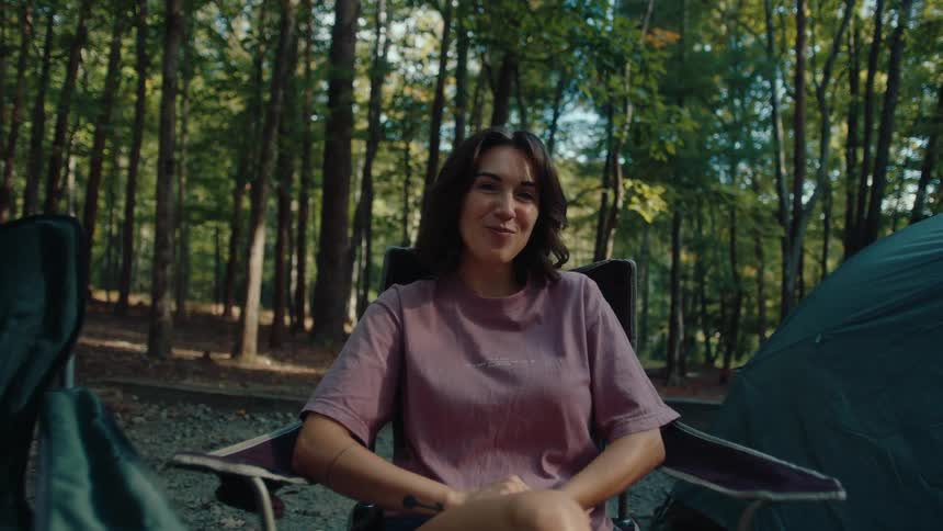 A young woman relaxes in a camp chair at her forested campsite, smiling warmly at the camera as it zooms in