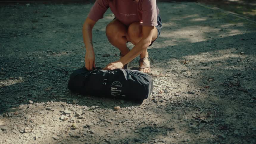 A woman sets down her tent bag and begins to unpack it at a sunny gravel campsite